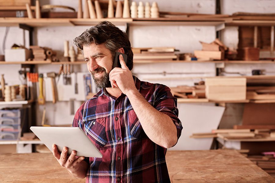 Contact - View of Smiling Business Owner Standing in His Woodworking Shop While Using a Tablet and Making a Call on His Phone